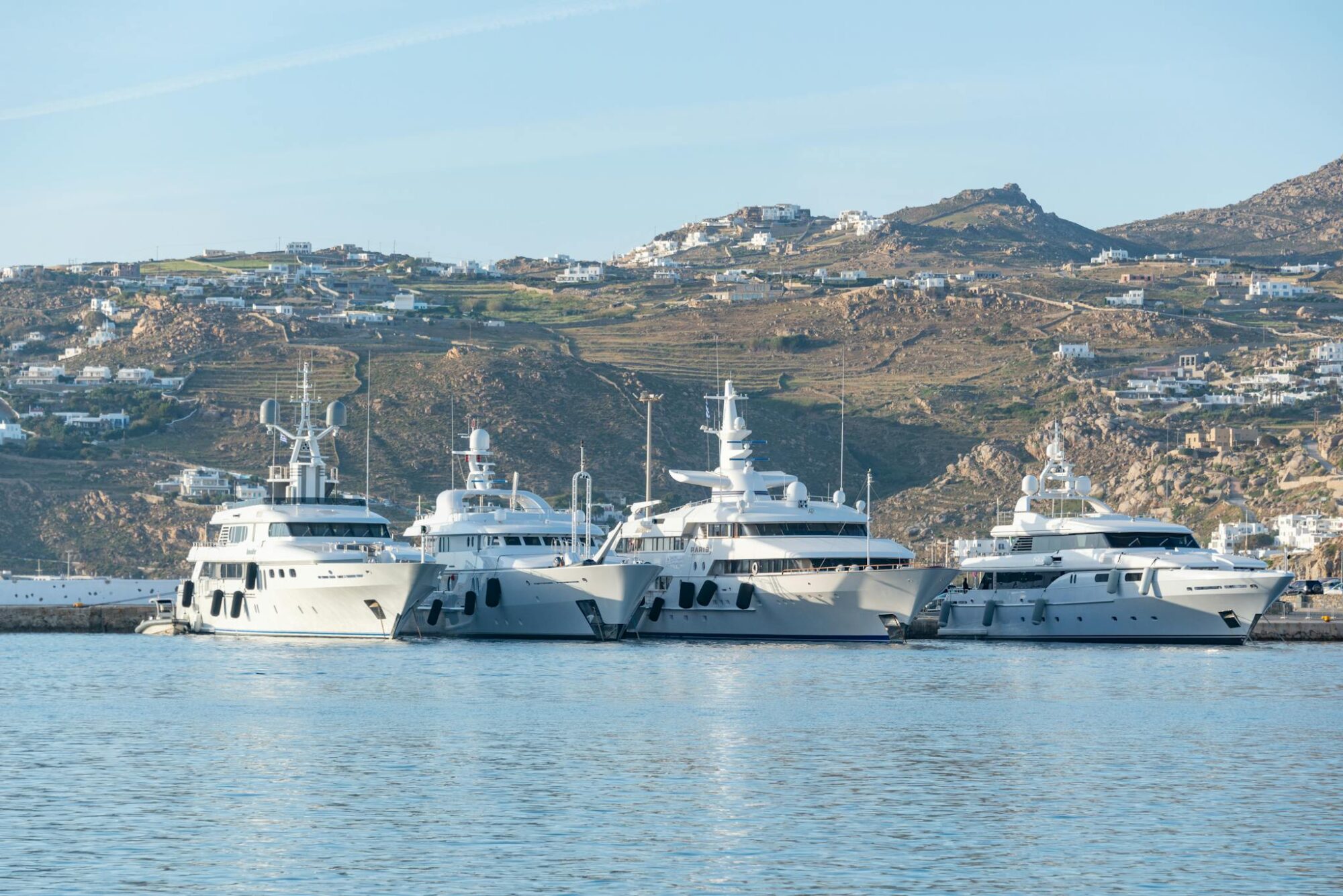 A row of luxury yachts moored at a picturesque harbor with hills in the background.